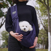 Person holding a small white dog in a purple sling with a blurred outdoor background