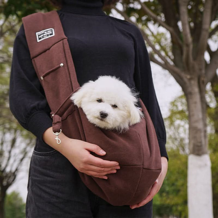 Person holding a small white dog in a brown sling bag outdoors.