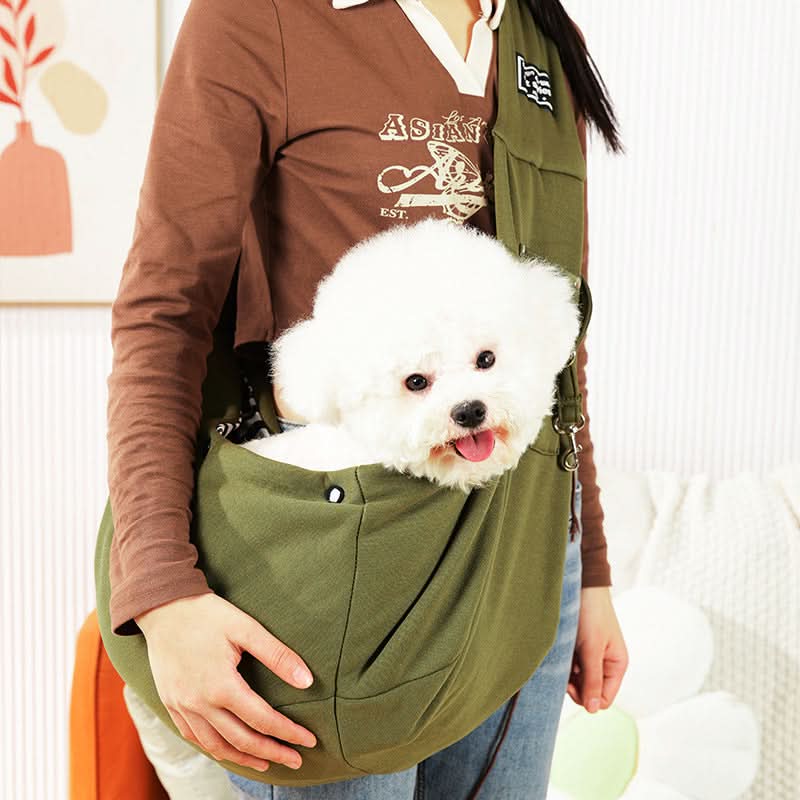 Person holding a white dog in an olive green pet sling against a neutral background