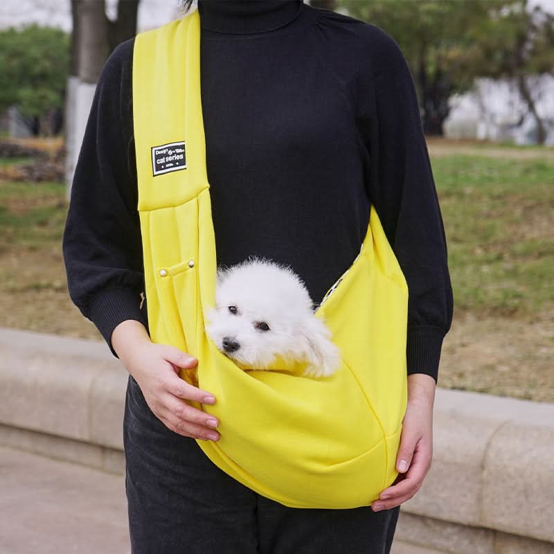 Person holding a small white dog in a yellow sling outdoors.