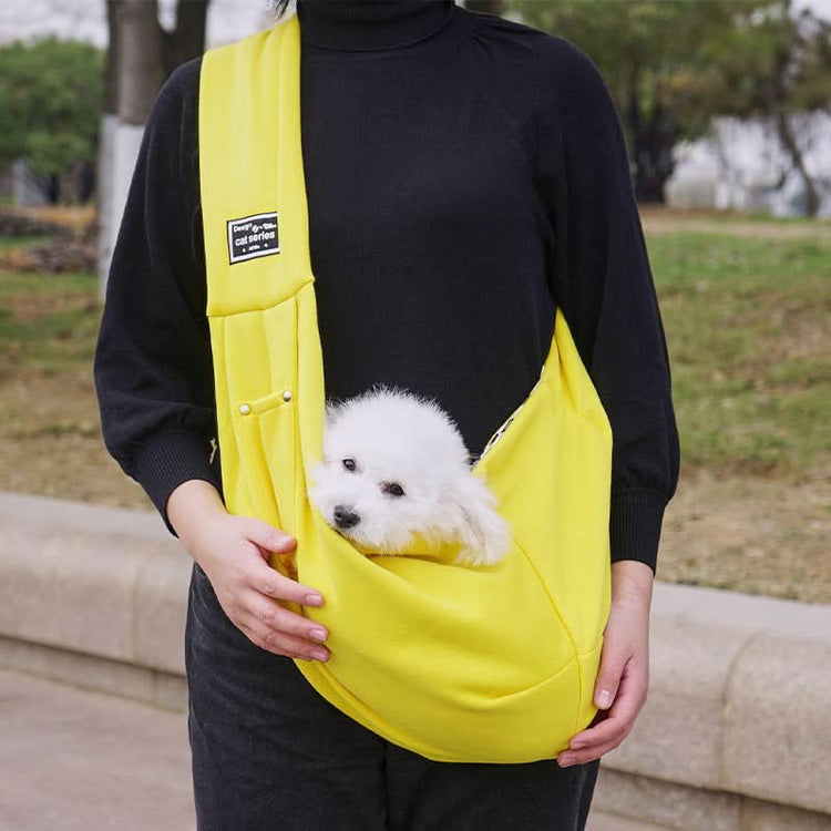 Person holding a small white dog in a yellow sling outdoors.