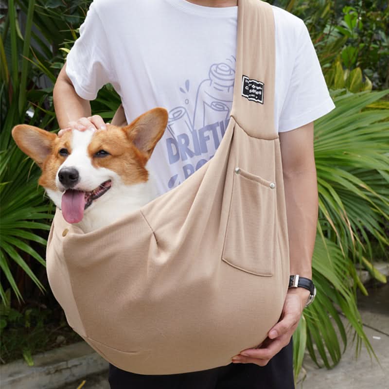 Person holding a corgi in a beige sling with green foliage in the background