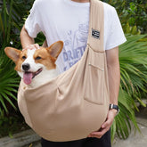 Person holding a corgi in a beige sling with green foliage in the background