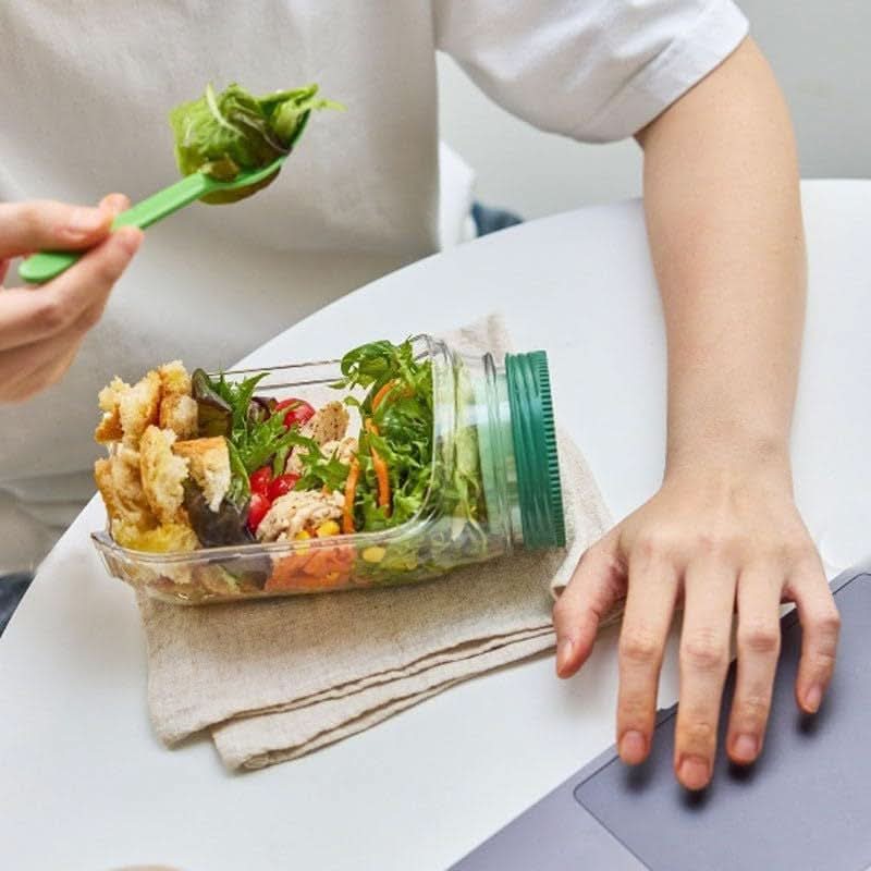 Person holding a jar of salad with a fork, sitting at a table.
