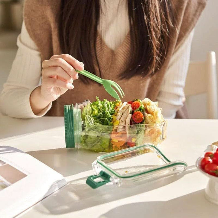 Person eating a salad from a glass container with a green fork on a white table.